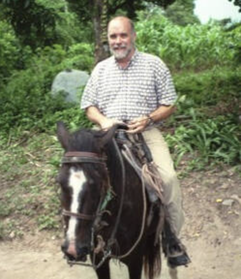 A man with a short beard and wearing a checked shirt is riding a dark brown horse. The background consists of lush greenery and a dirt path. The man seems relaxed and is holding the reins of the horse with both hands.